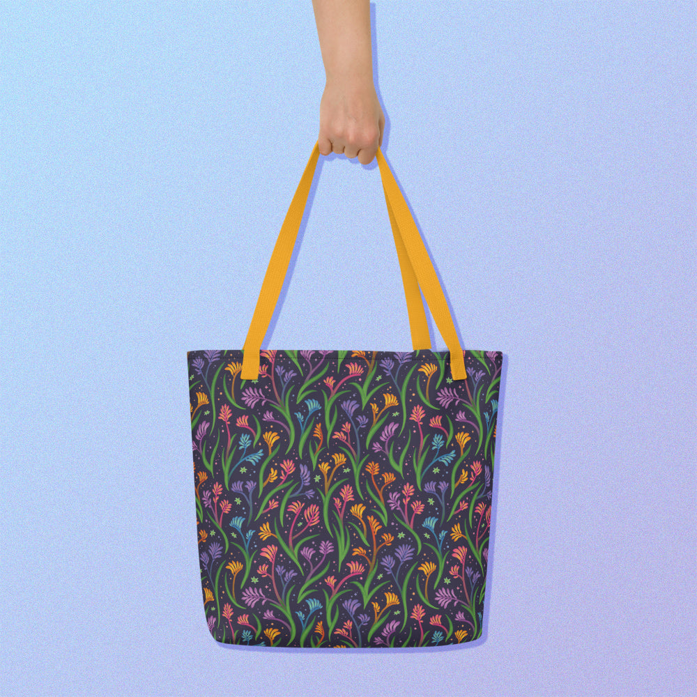 Hand holding a tote bag with a yellow strap and a floral rainbow kangaroo paw pattern on a gradient blue and purple background.