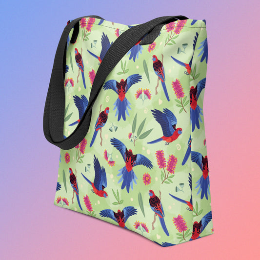 Tote bag with black handles and a crimson rosella parrot pattern on an angle on a gradient purple and pink background