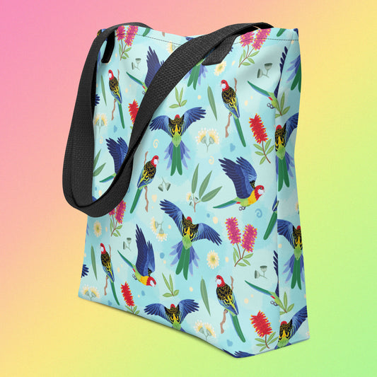 Tote bag with black handles and an eastern rosella parrot pattern on an angle on a gradient pink, yellow and green background