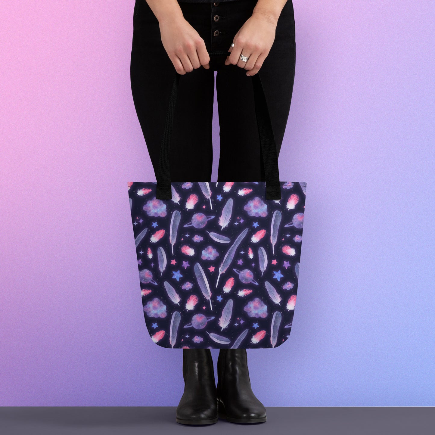 Person holding a tote bag with a black strap and galah cockatoo feather and galaxy-themed pattern on a gradient purple background