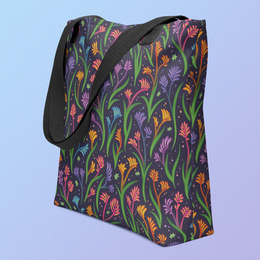 Tote bag on an angle with a black strap and a floral rainbow kangaroo paw pattern on a gradient blue and purple background.