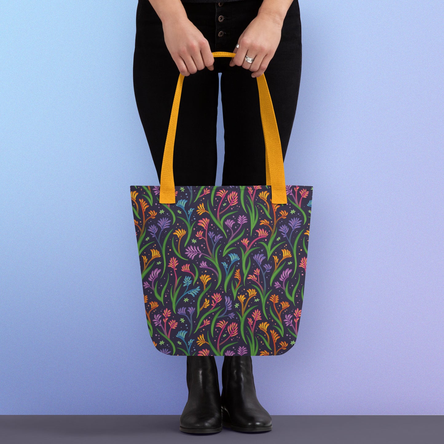 Person holding a tote bag with a yellow strap and a floral rainbow kangaroo paw pattern on a gradient blue and purple background.