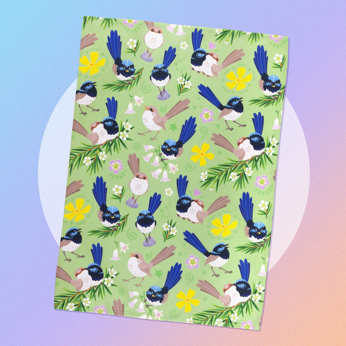A tea towel with a green, blue, yellow and beige superb fairywrens pattern sitting on a white circle on a pastel blue, purple and apricot background.