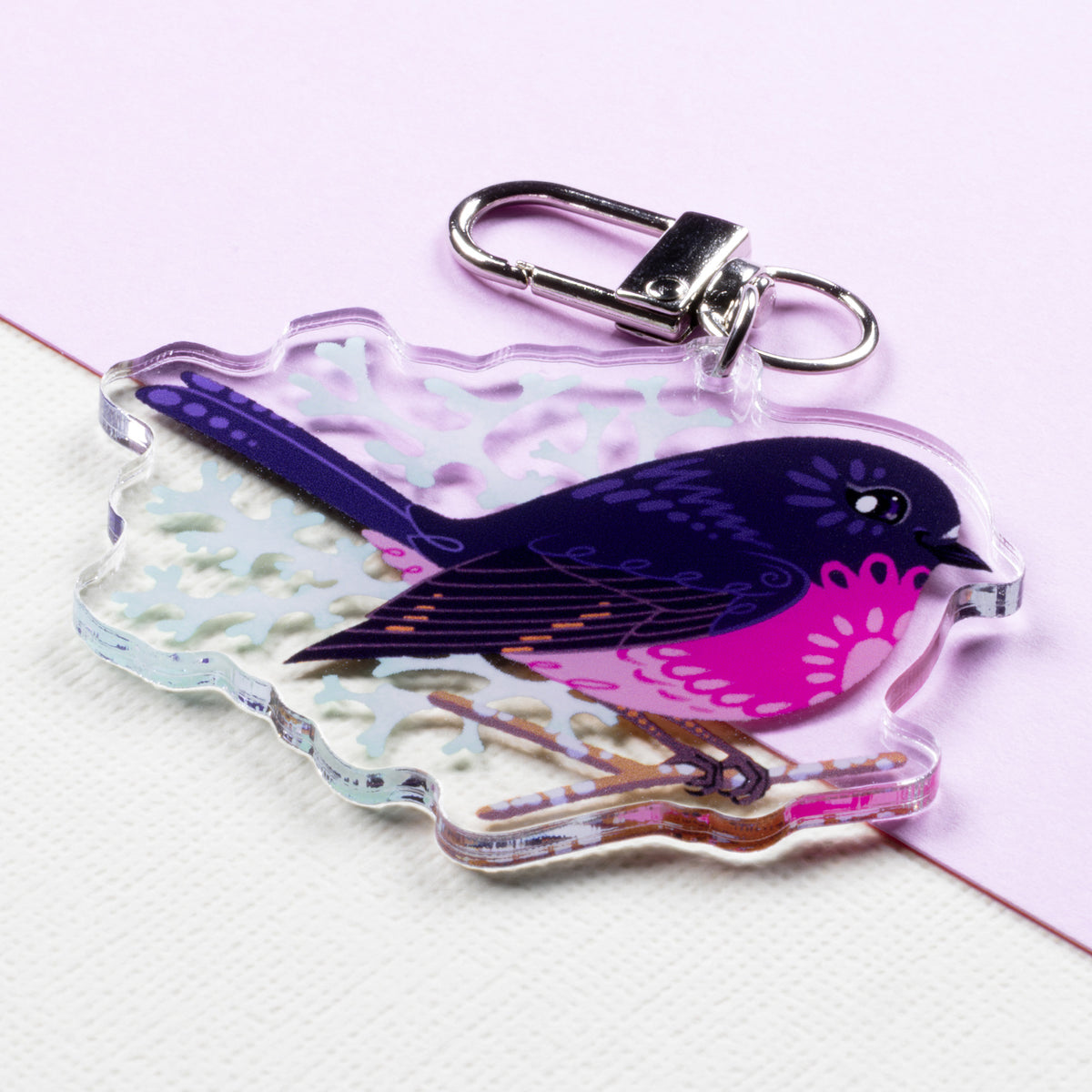 Angled photo of a double-sided acrylic keychain of male & female pink robins sitting on a split cream and pale lilac background.