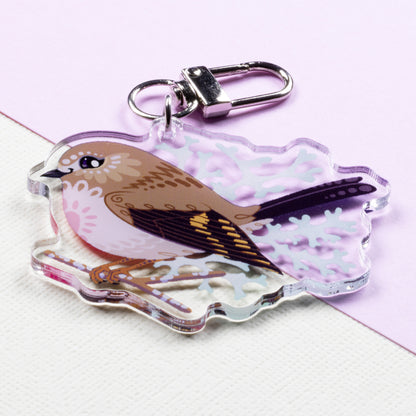 Angled photo of a double-sided acrylic keychain of male & female pink robins sitting on a split cream and pale lilac background.