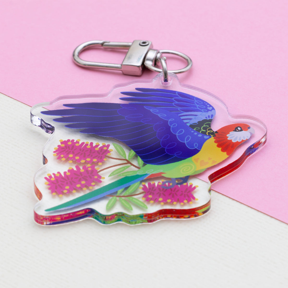 Angled photo of a double-sided acrylic keychain of crimson & eastern rosellas sitting on a split cream and pale pink background.