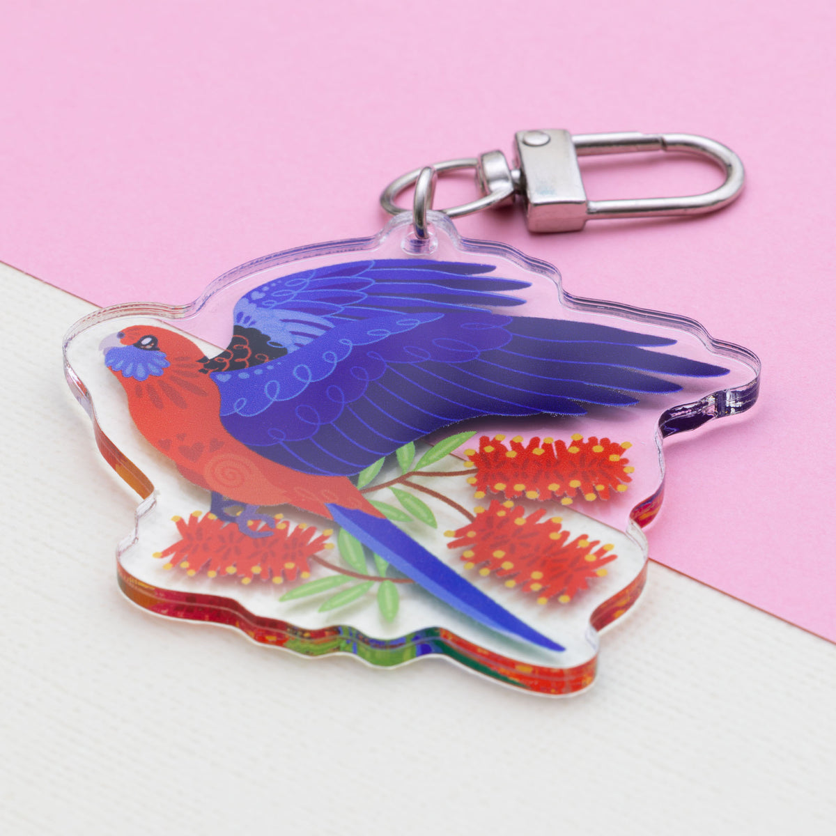 Angled photo of a double-sided acrylic keychain of crimson & eastern rosellas sitting on a split cream and pale pink background.