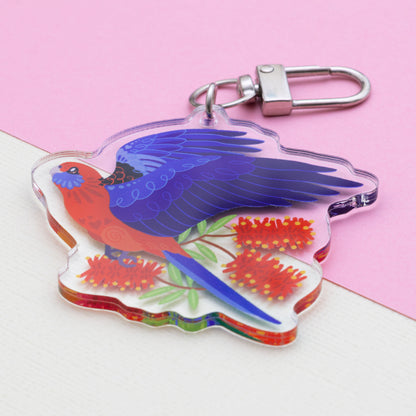 Angled photo of a double-sided acrylic keychain of crimson & eastern rosellas sitting on a split cream and pale pink background.