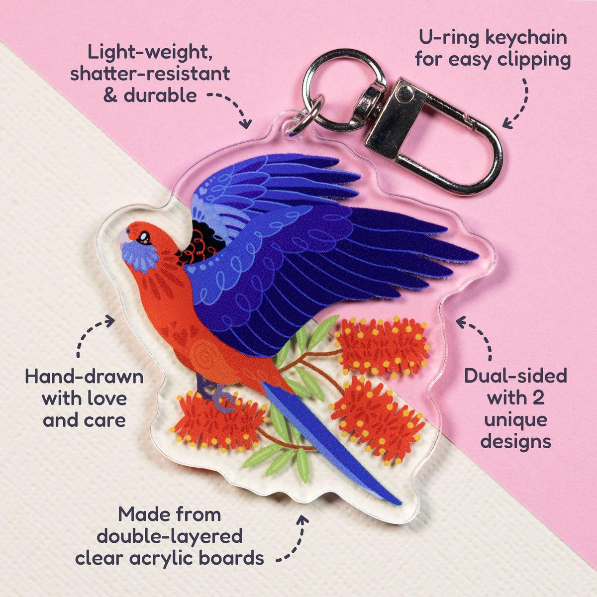Features & benefits (U-ring keychain for easy clipping, light-weight, shatter-resistant & durable, hand-drawn with love and care, made from double-layered clear acrylic boards, dual-sided with 2 unique designs) surrounding a double-sided acrylic keychain of crimson & eastern rosellas sitting on a split cream and pale pink background.