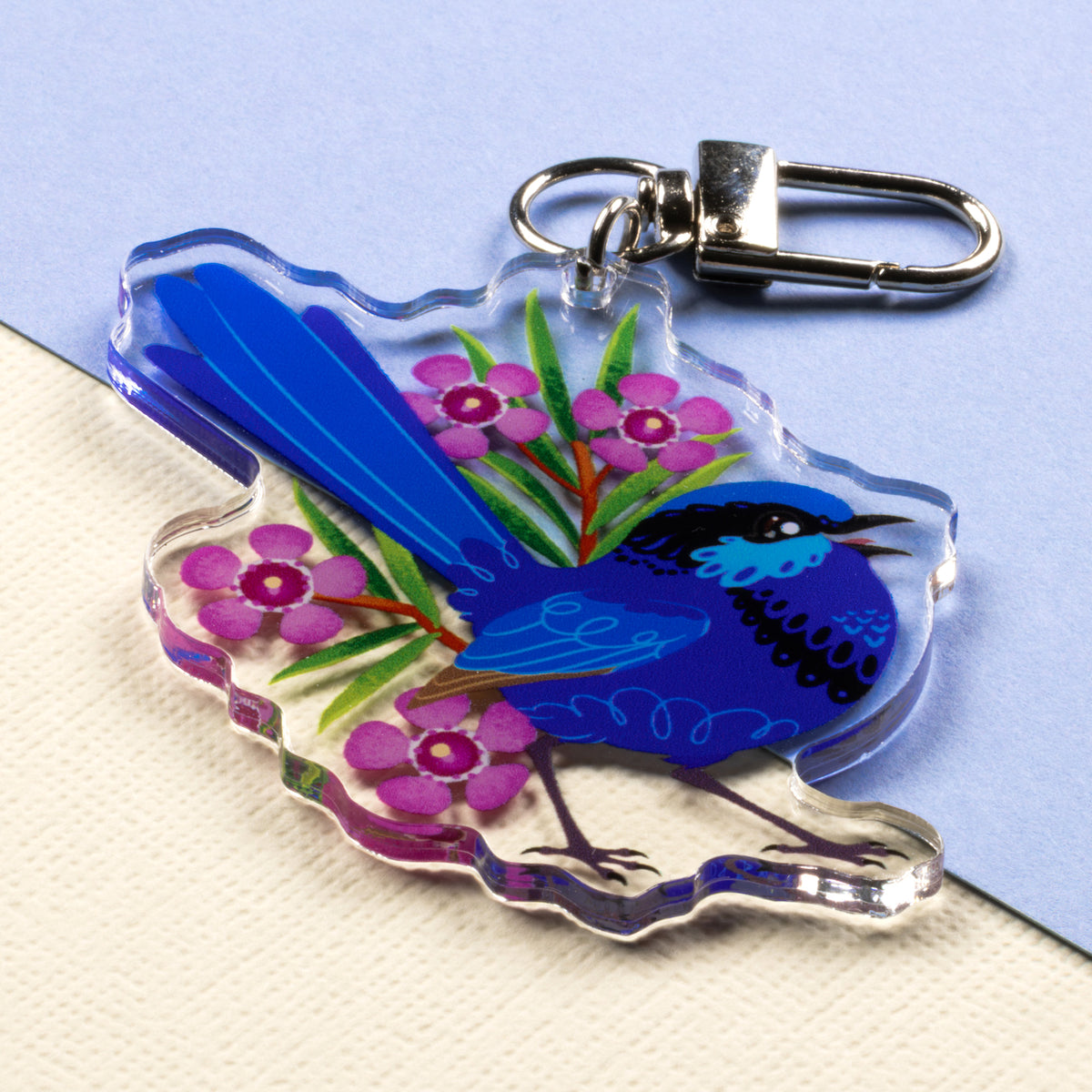 Angled photo of a double-sided acrylic keychain of male & female splendid fairywrens sitting on a split cream and pale blue background.