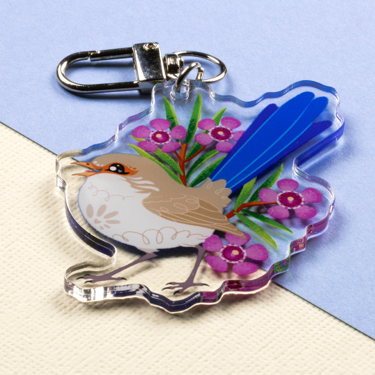 Angled photo of a double-sided acrylic keychain of male & female splendid fairywrens sitting on a split cream and pale blue background.