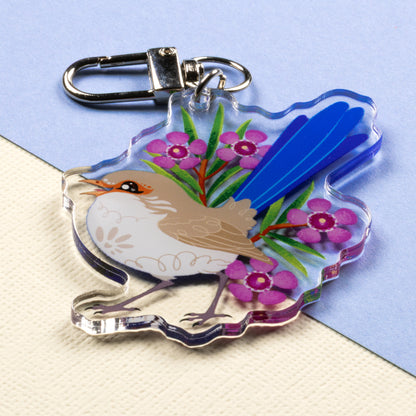 Angled photo of a double-sided acrylic keychain of male & female splendid fairywrens sitting on a split cream and pale blue background.