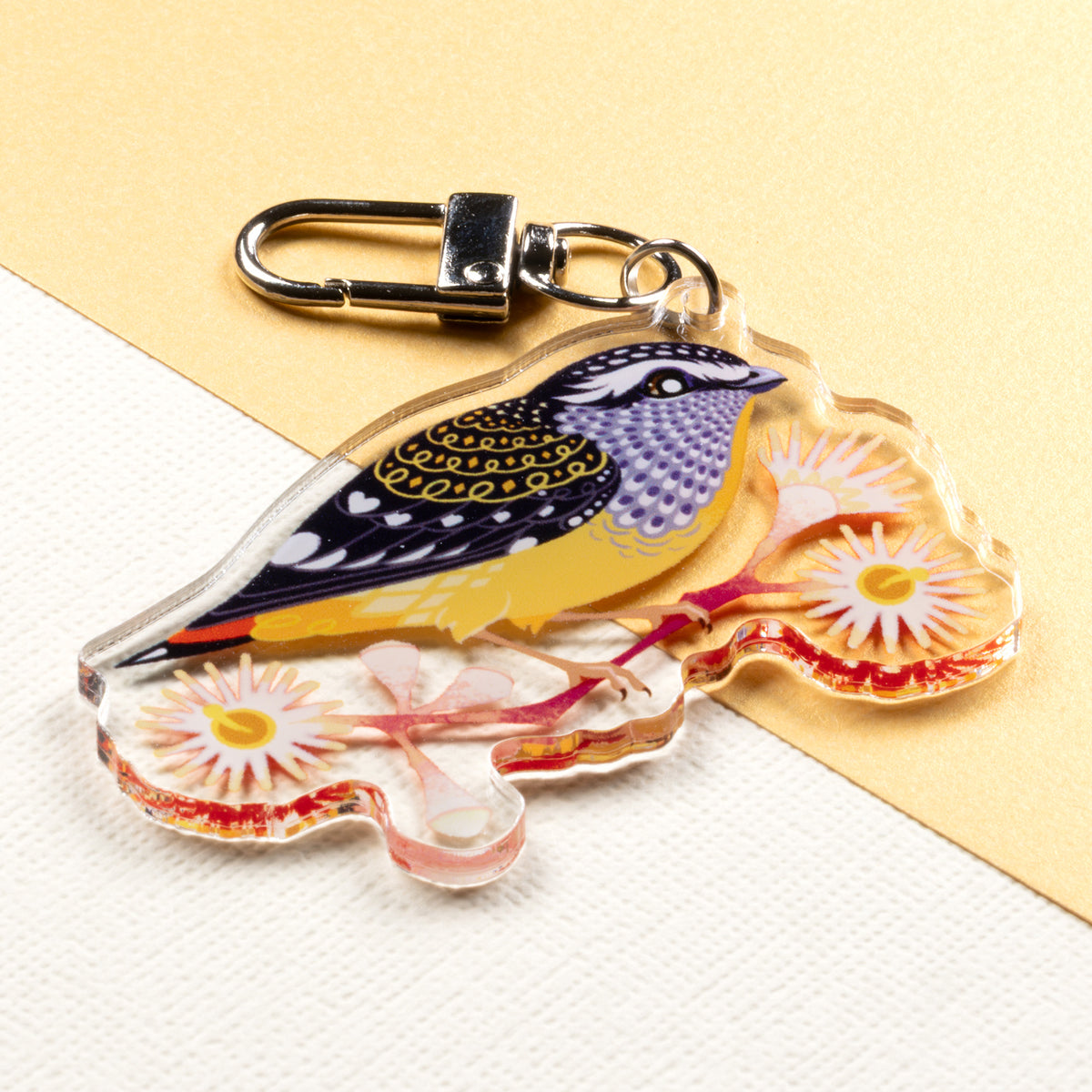 Angled photo of a double-sided acrylic keychain of male & female spotted pardalotes sitting on a split cream & metallic gold background. 