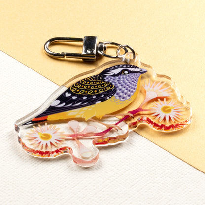 Angled photo of a double-sided acrylic keychain of male & female spotted pardalotes sitting on a split cream & metallic gold background. 