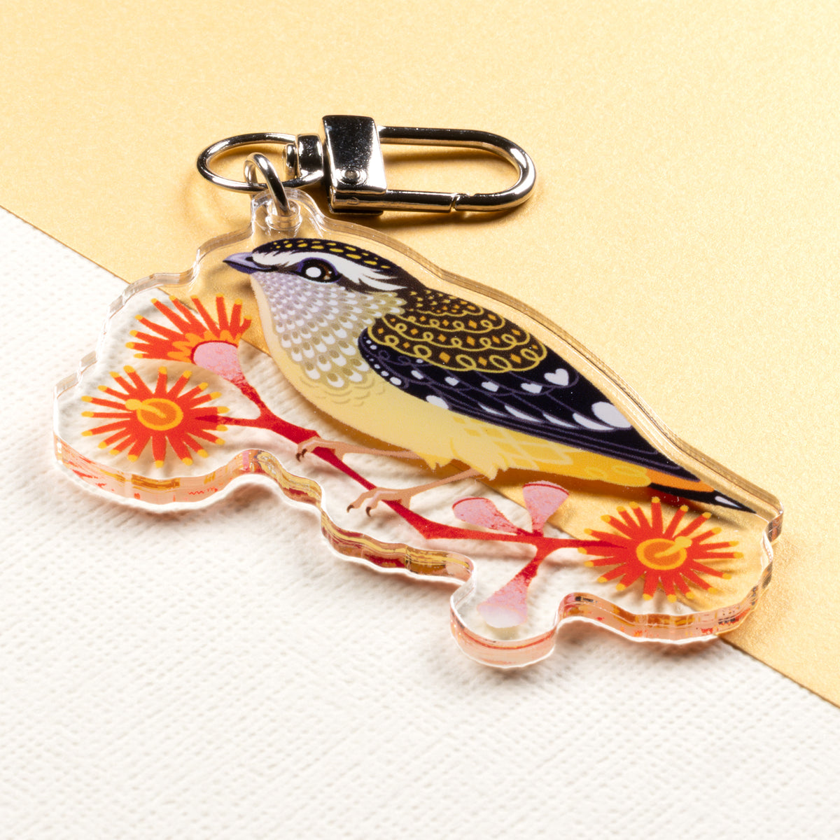 Angled photo of a double-sided acrylic keychain of male & female spotted pardalotes sitting on a split cream & metallic gold background. 