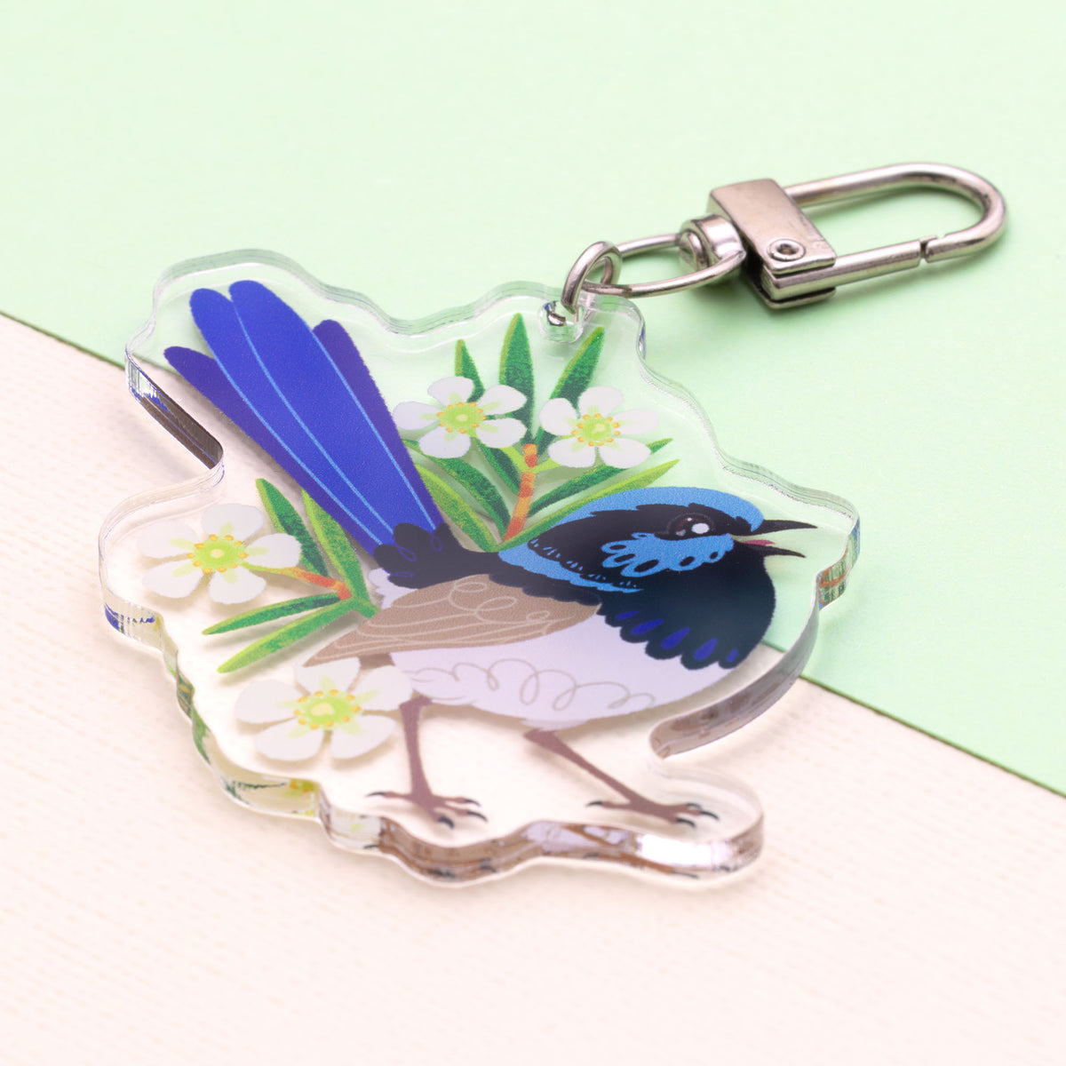 Angled photo of a double-sided acrylic keychain of a male and female superb fairywren sitting on a split cream and pale green background.