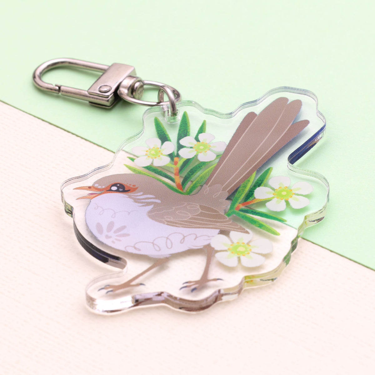 Angled photo of a double-sided acrylic keychain of a male and female superb fairywren sitting on a split cream and pale green background.
