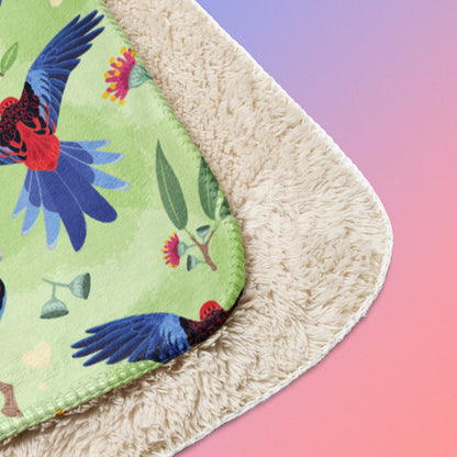 Close up detail of folded green, red and blue crimson rosella & native Aussie plant pattern blanket on a blue and pink gradient background.