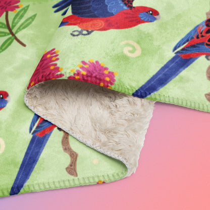 Close up detail of folded green, red and blue crimson rosella & native Aussie plant pattern blanket on a blue and pink gradient background.