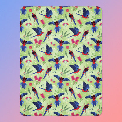 A green, red and blue crimson rosella & native Aussie plant pattern blanket on a blue and pink gradient background.