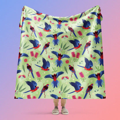 Person holding a green, red and blue crimson rosella & native Aussie plant pattern blanket above their head on a blue and pink gradient background.