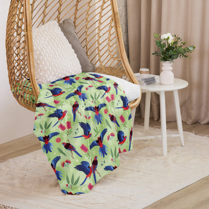 A folded green, red and blue crimson rosella & native Aussie plant pattern blanket sitting on a swinging woven egg chair with soft, natural lighting.