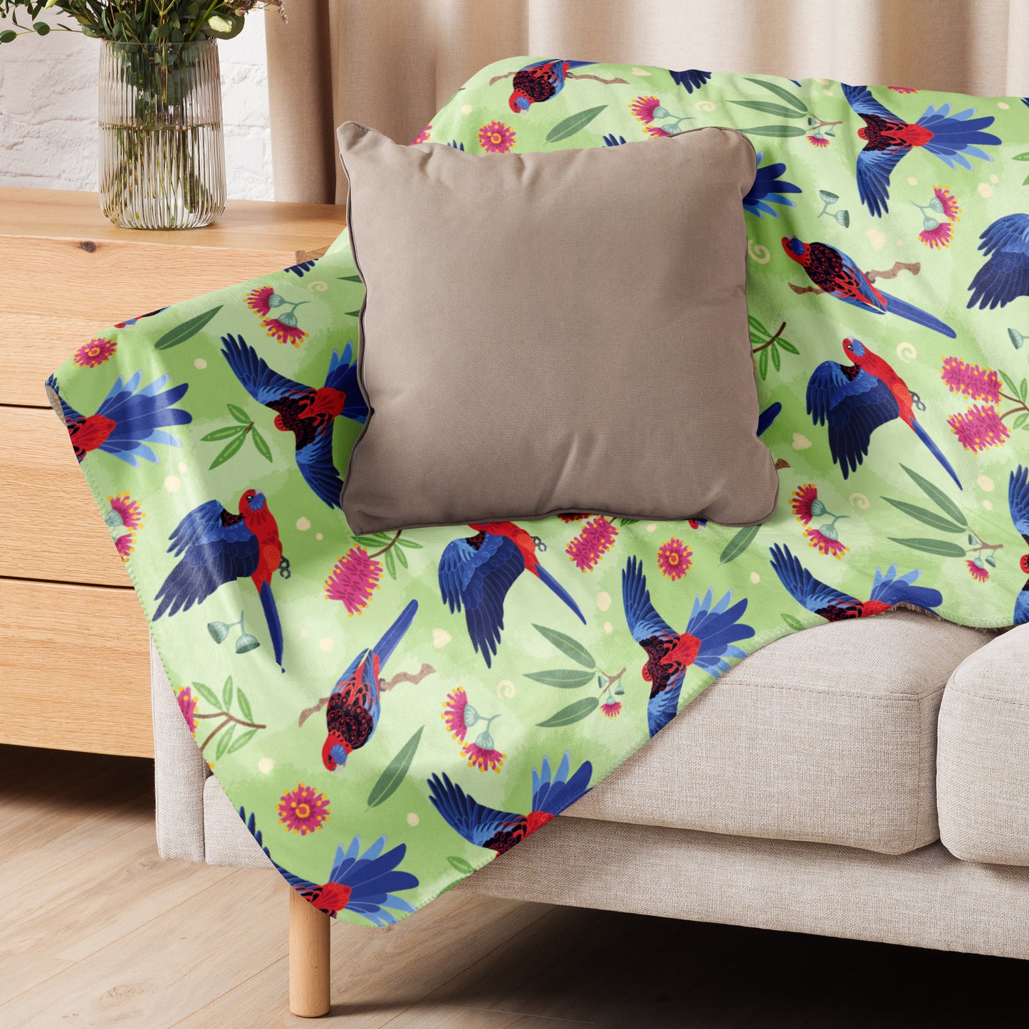 A folded green, red and blue crimson rosella & native Aussie plant pattern blanket sitting on a beige sofa with a beige pillow on top.