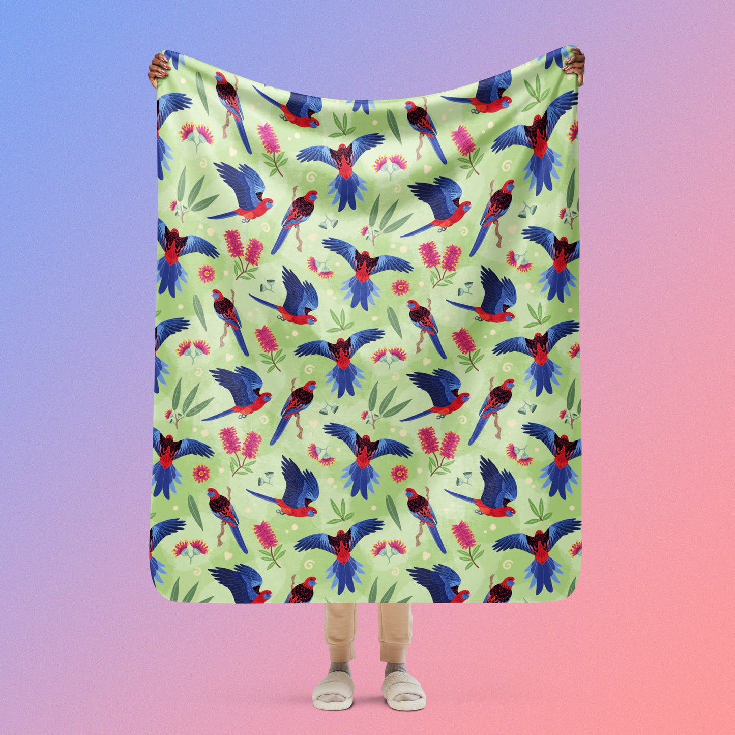 Person holding a green, red and blue crimson rosella & native Aussie plant pattern blanket above their head on a blue and pink gradient background.