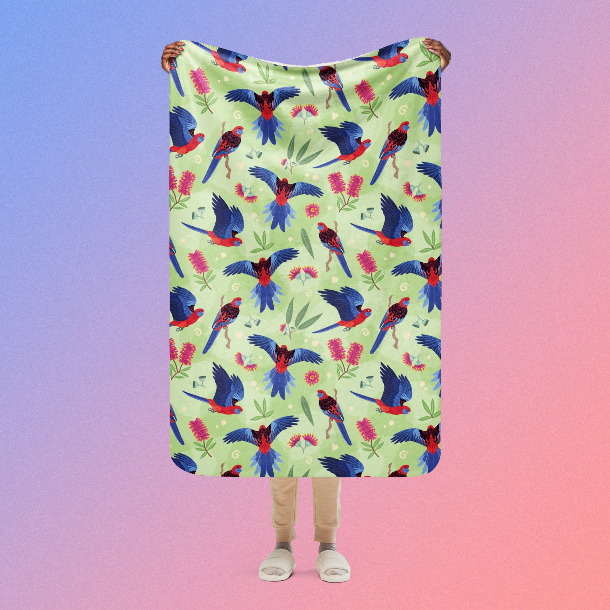 Person holding a green, red and blue crimson rosella & native Aussie plant pattern blanket above their head on a blue and pink gradient background.