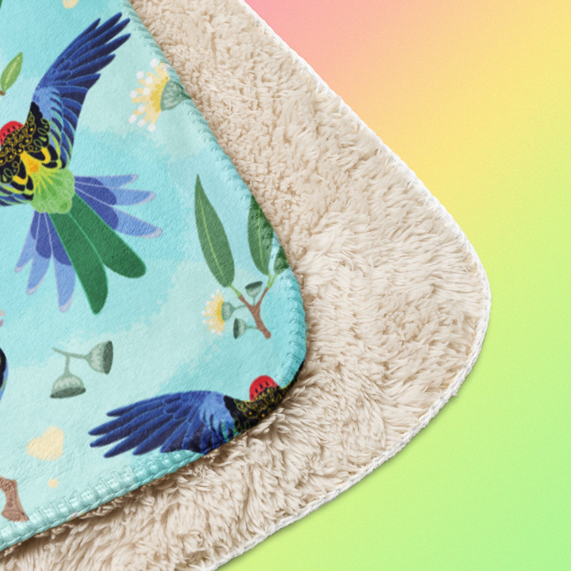 Close up detail of a folded blue, pink and green eastern rosella & native Aussie plant pattern blanket on a pink, yellow and green gradient background.