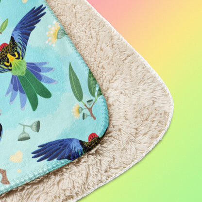 Close up detail of a folded blue, pink and green eastern rosella & native Aussie plant pattern blanket on a pink, yellow and green gradient background.