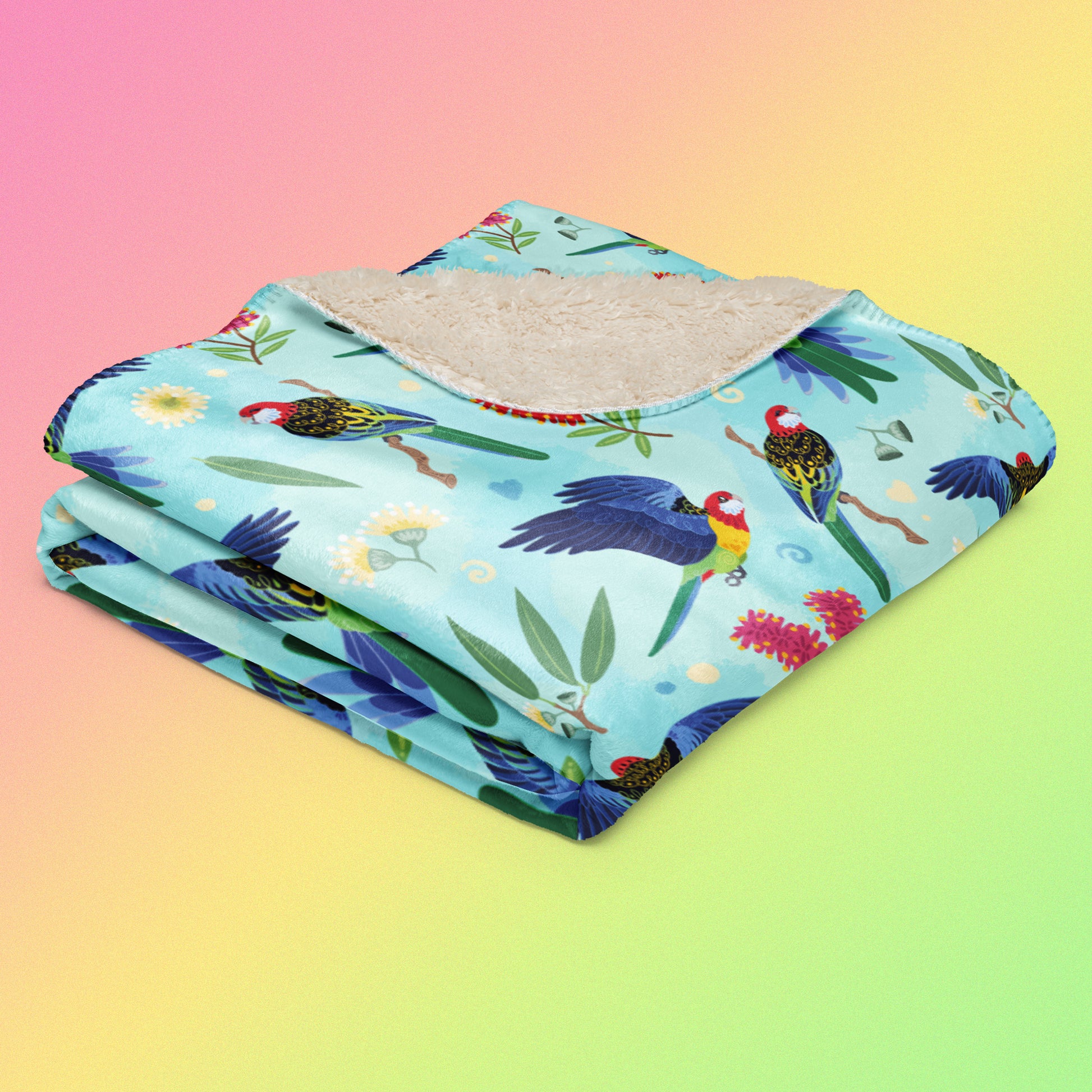 A folded blue, pink and green eastern rosella & native Aussie plant pattern blanket on a pink, yellow and green gradient background.