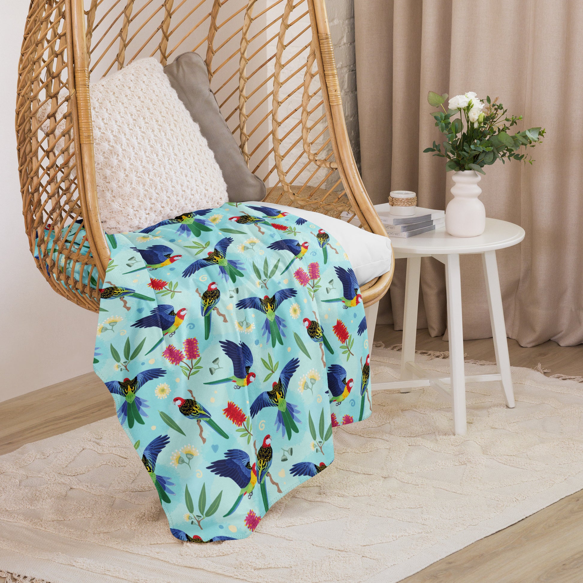 A folded blue, pink and green eastern rosella & native Aussie plant pattern blanket sitting on a swinging woven egg chair with soft, natural lighting.