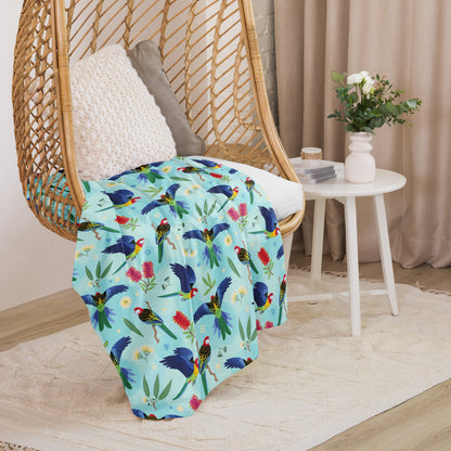 A folded blue, pink and green eastern rosella & native Aussie plant pattern blanket sitting on a swinging woven egg chair with soft, natural lighting.