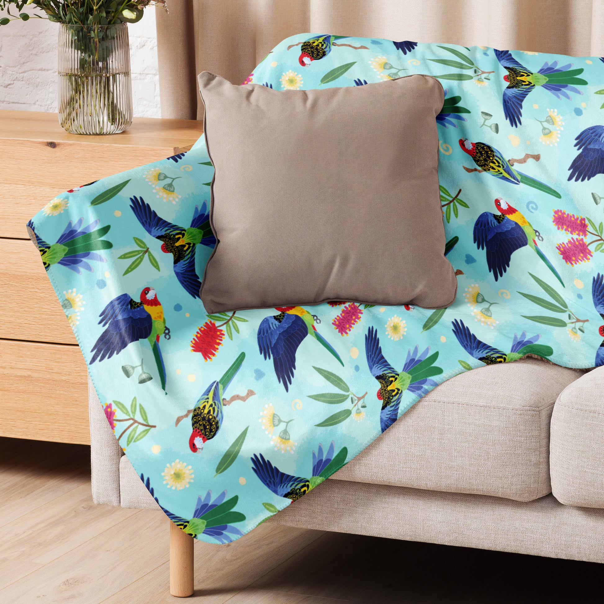 A folded blue, pink and green eastern rosella & native Aussie plant pattern blanket sitting on a beige sofa with a beige pillow on top.