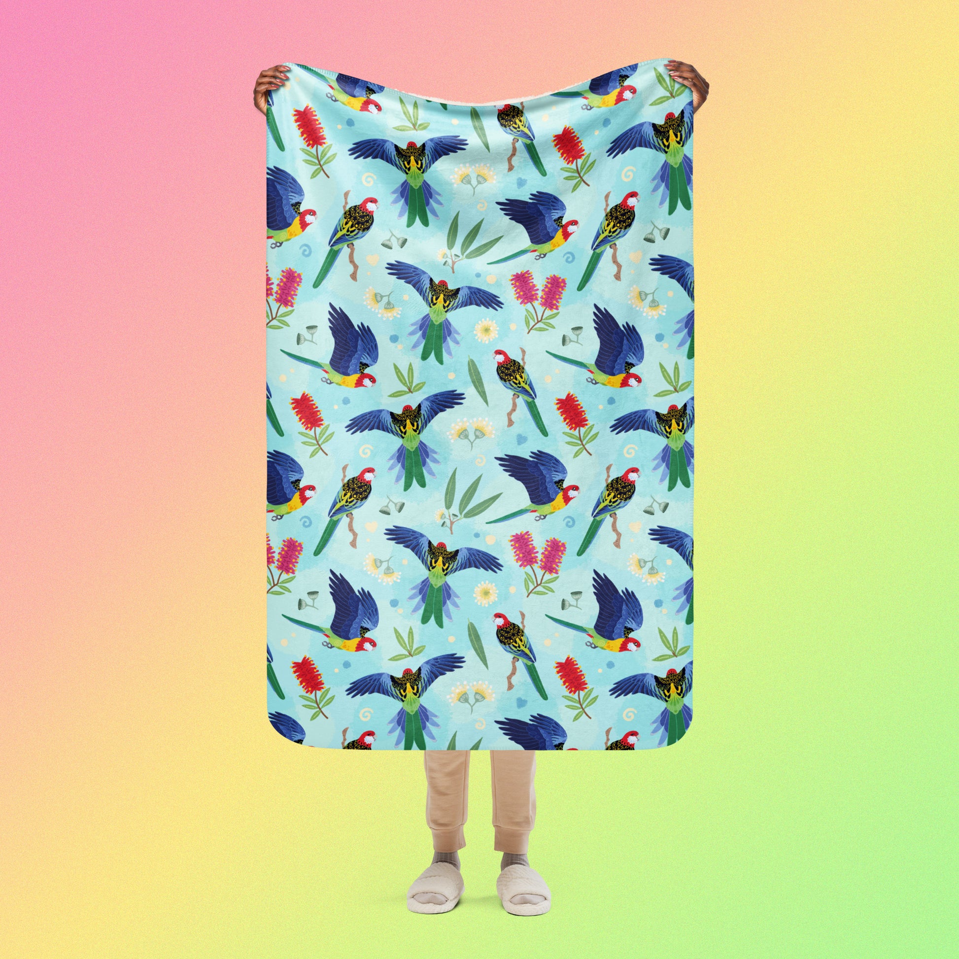 Person holding a blue, pink and green eastern rosella & native Aussie plant pattern blanket above their head on a pink, yellow and green gradient background.