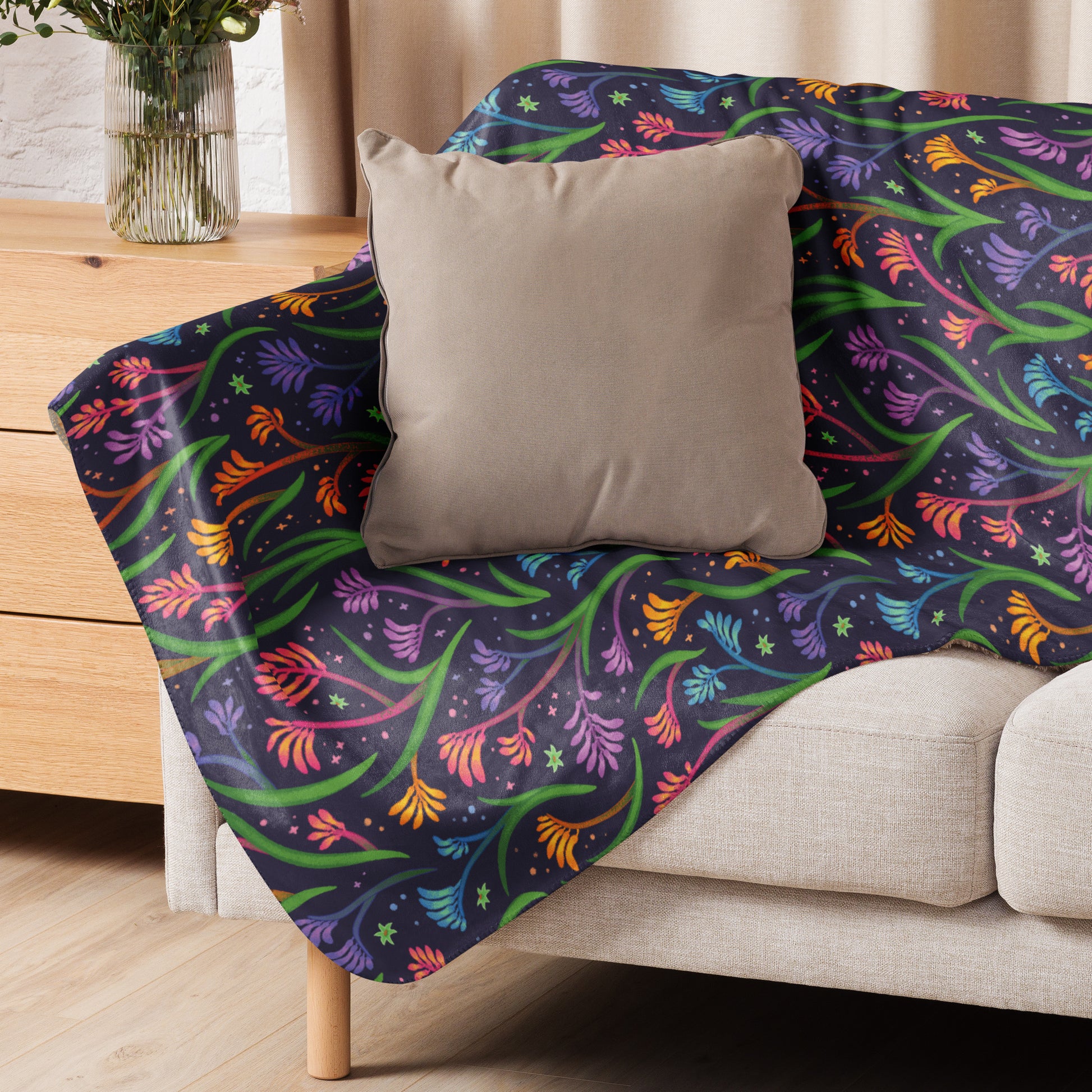 A rainbow kangaroo paw flower pattern blanket sitting on a beige sofa with a beige pillow on top.