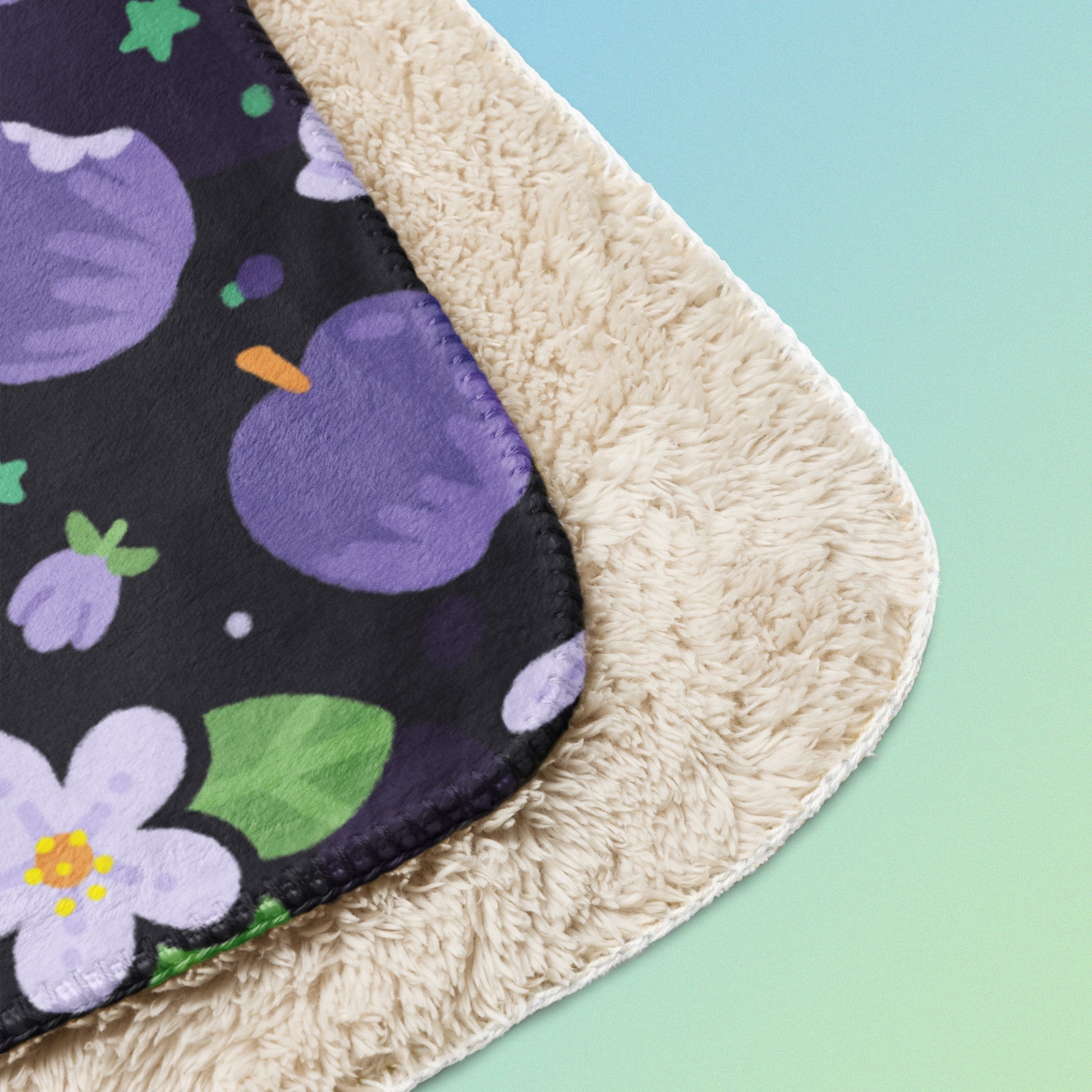 Close up detail of a folded purple apples & blossoms pattern blanket on a blue and green gradient background.