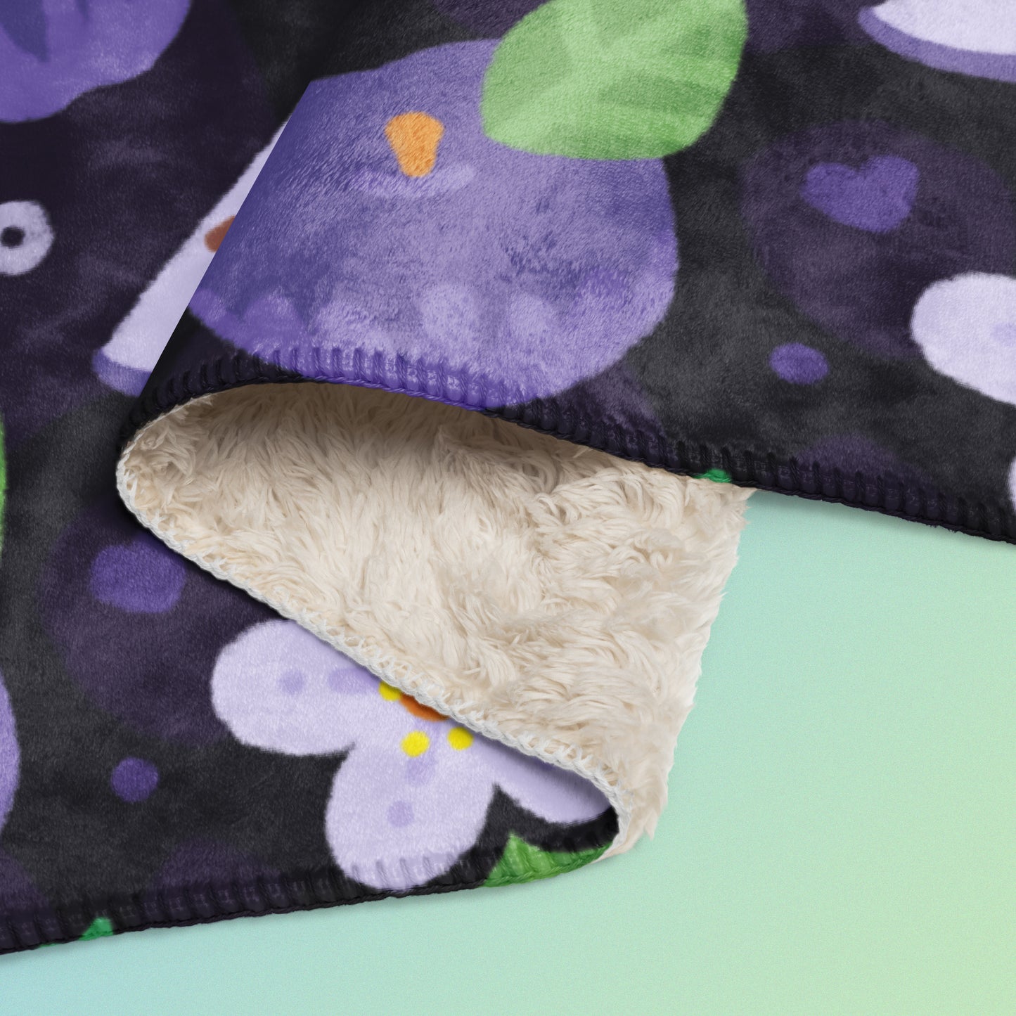 Close up detail of a folded purple apples & blossoms pattern blanket on a blue and green gradient background.