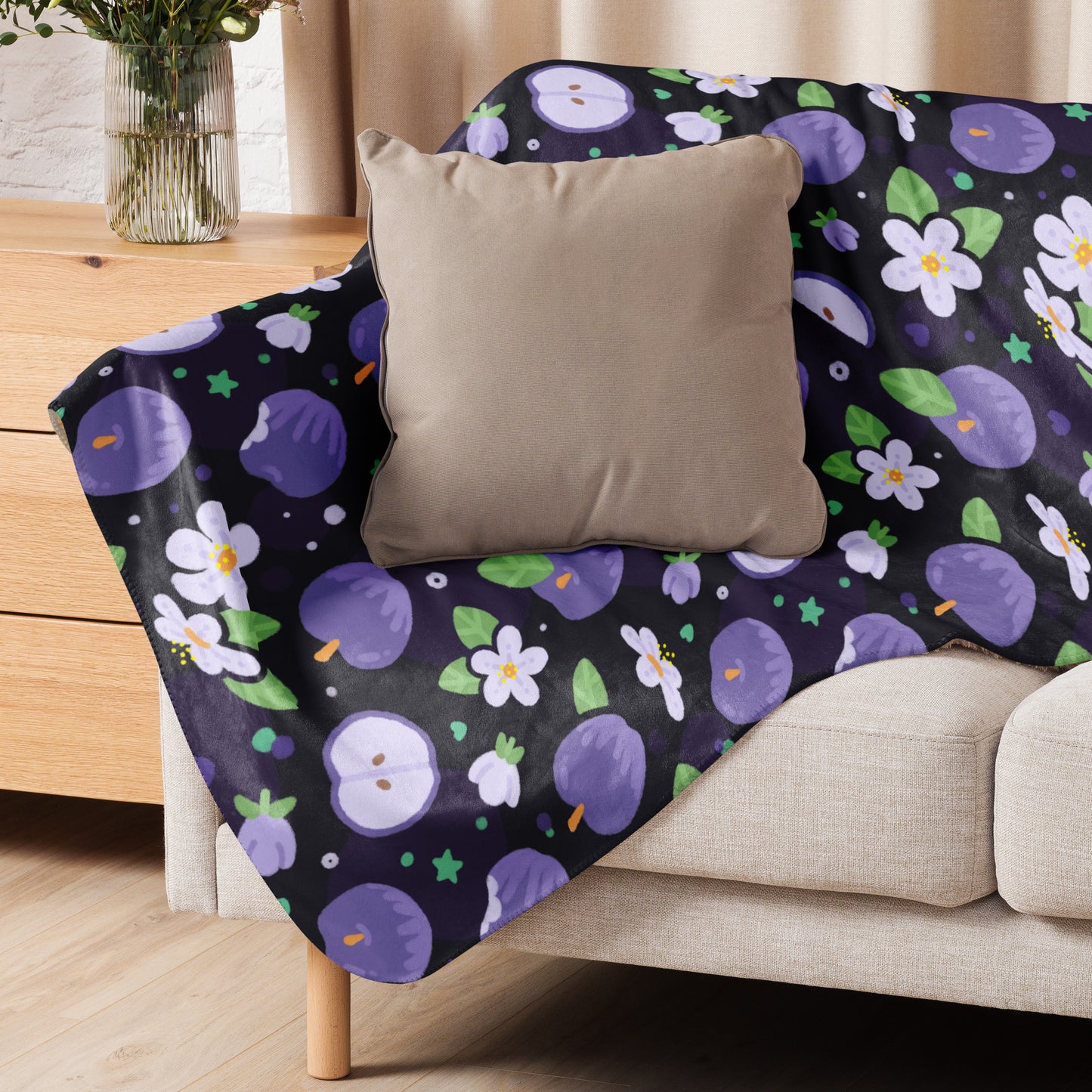 A purple apples & blossoms pattern blanket sitting on a beige sofa with a beige pillow on top.