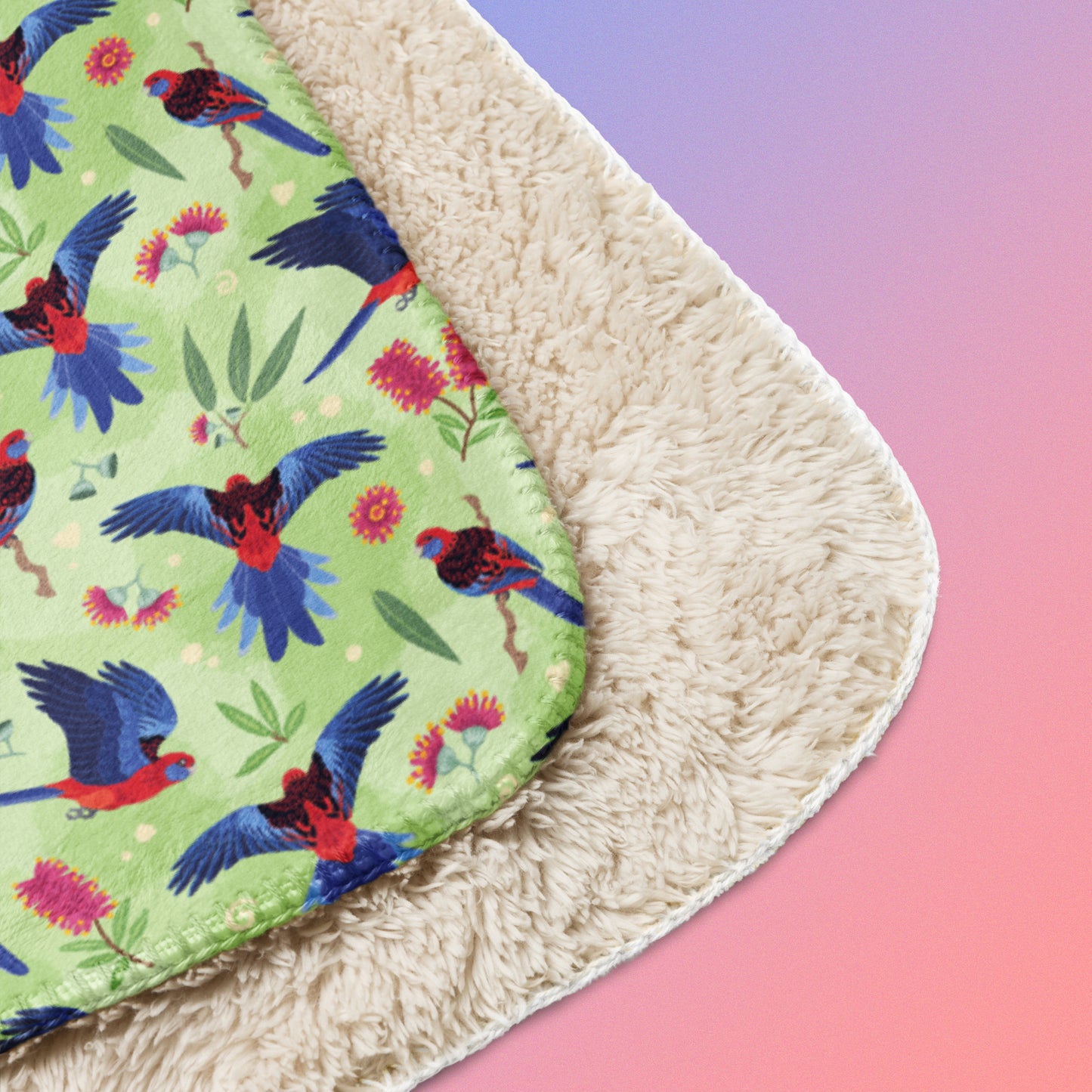Close up detail of folded green, red and blue crimson rosella & native Aussie plant pattern blanket on a blue and pink gradient background.