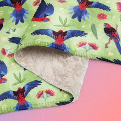 Close up detail of folded green, red and blue crimson rosella & native Aussie plant pattern blanket on a blue and pink gradient background.