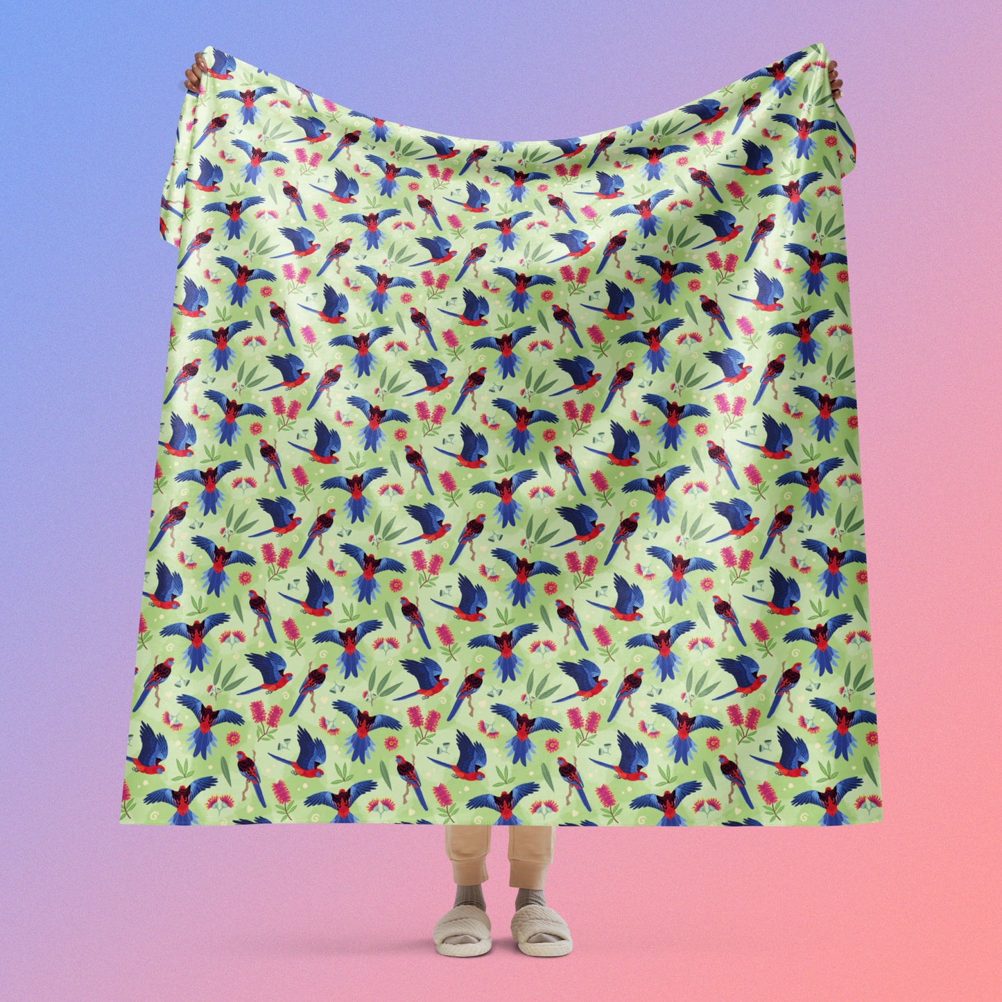 Person holding a green, red and blue crimson rosella & native Aussie plant pattern blanket above their head on a blue and pink gradient background.