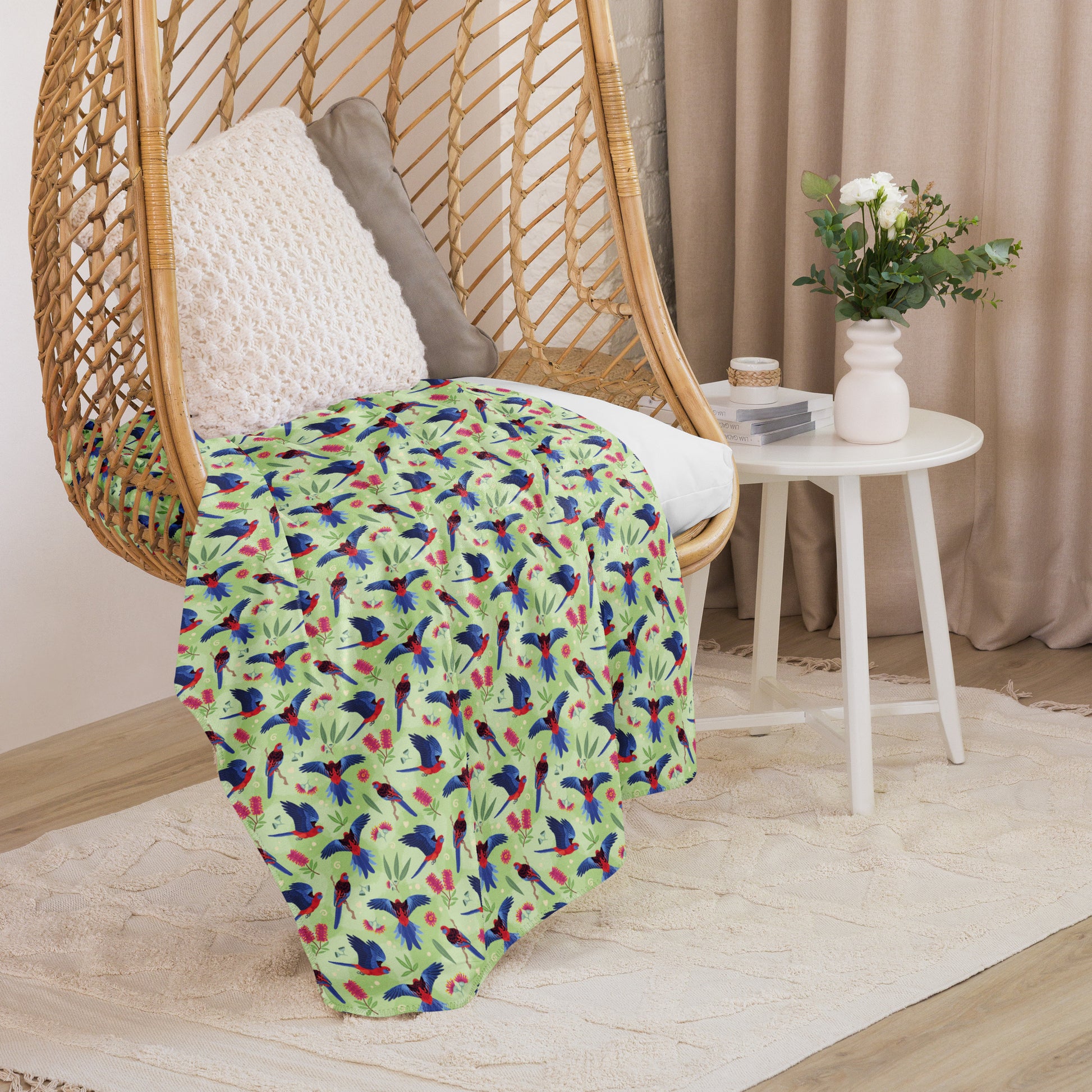 A folded green, red and blue crimson rosella & native Aussie plant pattern blanket sitting on a swinging woven egg chair with soft, natural lighting.