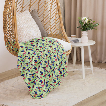 A folded green, red and blue crimson rosella & native Aussie plant pattern blanket sitting on a swinging woven egg chair with soft, natural lighting.