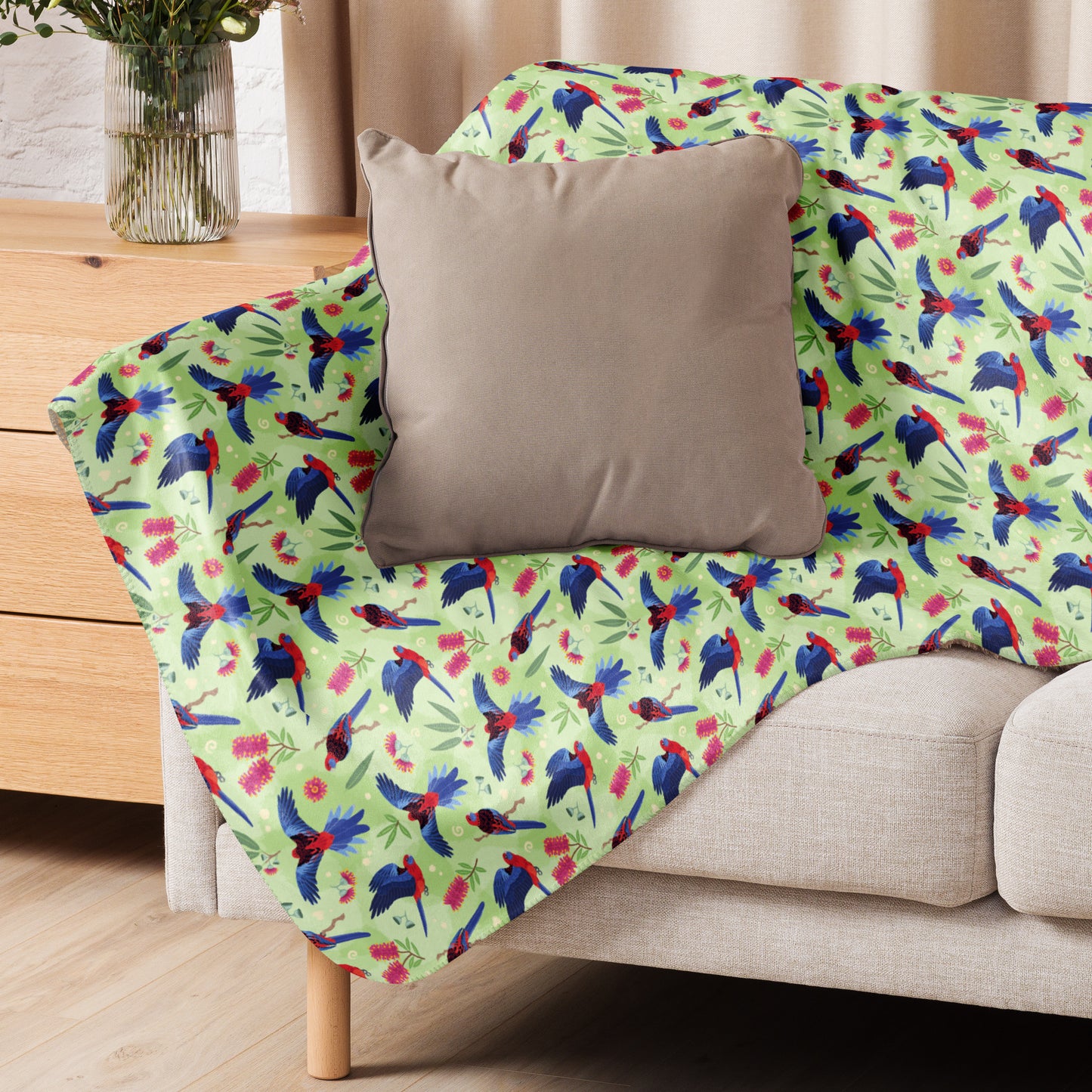 A folded green, red and blue crimson rosella & native Aussie plant pattern blanket sitting on a beige sofa with a beige pillow on top.