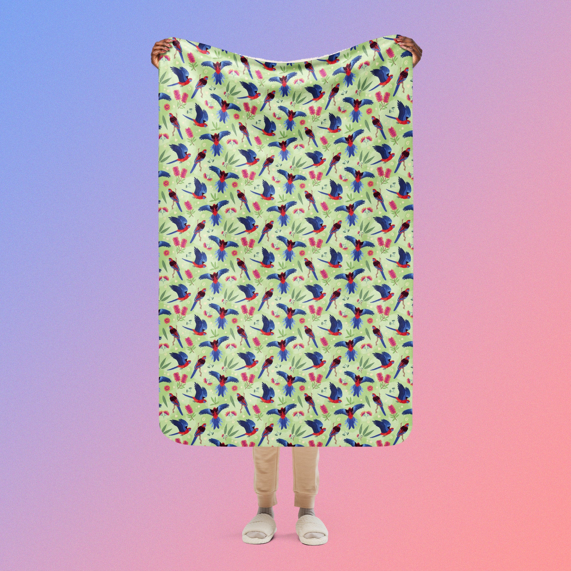 Person holding a green, red and blue crimson rosella & native Aussie plant pattern blanket above their head on a blue and pink gradient background.
