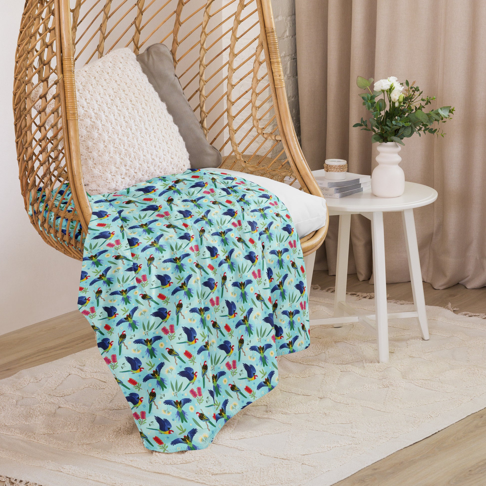 A folded blue, pink and green eastern rosella & native Aussie plant pattern blanket sitting on a swinging woven egg chair with soft, natural lighting.