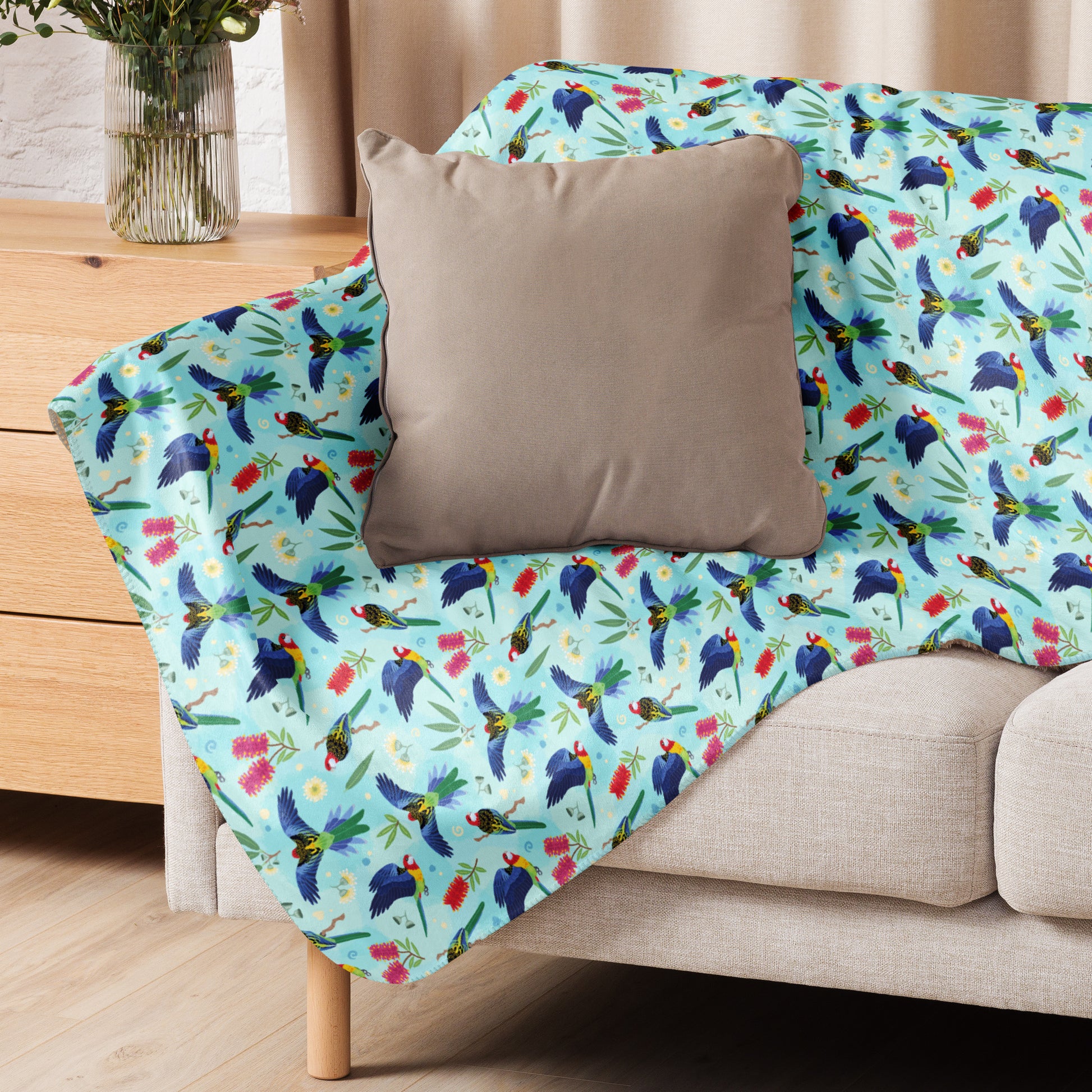 A folded blue, pink and green eastern rosella & native Aussie plant pattern blanket sitting on a beige sofa with a beige pillow on top.