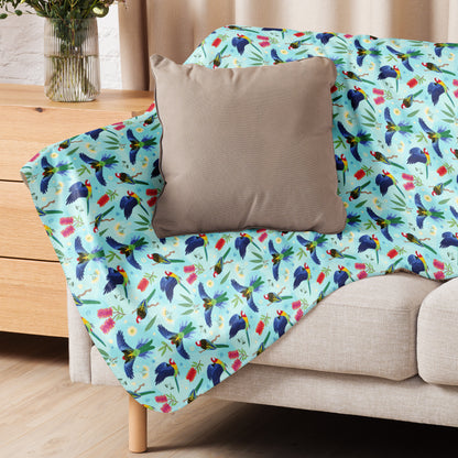 A folded blue, pink and green eastern rosella & native Aussie plant pattern blanket sitting on a beige sofa with a beige pillow on top.